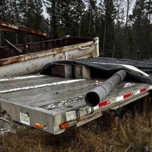 Rusty flatbed trailer with a large metal pipe resting on it, covered by a tarp, in a snowy, wooded lot.