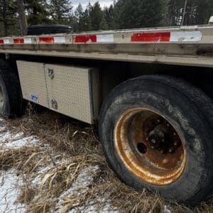 Close-up of a rusted trailer wheel and metal storage box beneath a flatbed trailer in a grassy, snowy area.