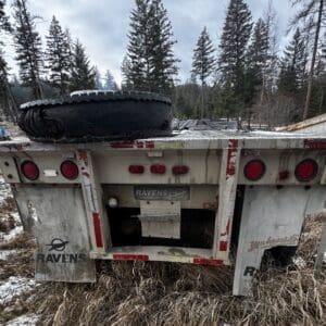 Rear view of a weathered Ravens trailer with a large spare tire mounted on top, parked in a snowy, grassy clearing among tall evergreen trees.