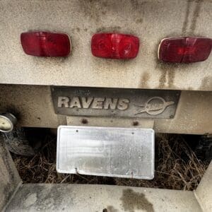 Rear of a dusty Ravens trailer with three red taillights, a metal 'RAVENS' badge, and an empty license plate mount.