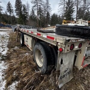 Flatbed trailer parked in a wintry field with tall pine trees in the background, showing a spare tire and equipment on board.