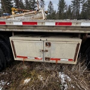 Rusted metal storage box under a trailer bed with red-and-white reflective strips, in a grassy, lightly snowy roadside area in a forested setting.