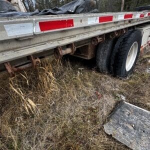 Flatbed trailer with rusted undercarriage and two tires, abandoned in dry grass with a wooden plank nearby.