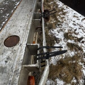 Close-up of a weathered trailer with black ratchet straps securing a metal bar along the side, snow and dry grass in the background.