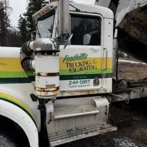 White truck cab with yellow/green stripe reading Foothills Trucking & Excavating; rust on exhaust and metal step area.
