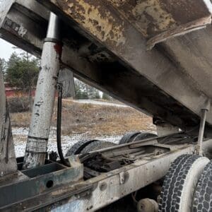 Close-up of an old, rusted dump truck with its bed raised by a hydraulic ram, showing a weathered frame and dual rear wheels in a snowy, rural lot.