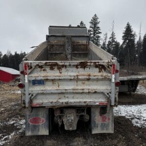 Rear view of a rusty white dump truck in a rural lot, with a rusted bed and muddy, snow-dotted ground and evergreen trees in the background on a cloudy day.