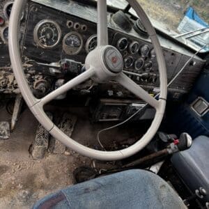 Old, dusty truck interior with a large white steering wheel and a weathered dashboard full of round gauges.