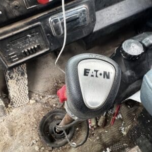 Close-up of a dusty Eaton gear shift lever in a dirty vehicle cab, with red knob and dashboard controls nearby.
