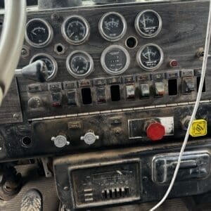 Vintage truck dashboard with round gauges (fuel, oil pressure, temperature) and a row of toggles, red emergency stop button, and dusty controls beyond a worn steering column.