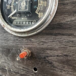Close-up of an old analog speedometer with the odometer, mounted on a worn wooden panel; a red push button sits nearby amid rust.