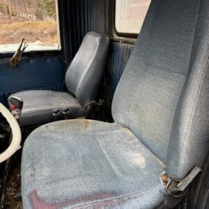Interior of an old blue truck cab with worn fabric seats, one torn, and dusty cabin surfaces visible through the window edge.