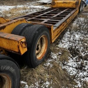 Rusty yellow trailer chassis with large worn tires on a snow-dusted field, a truck behind it and scattered equipment in the background.