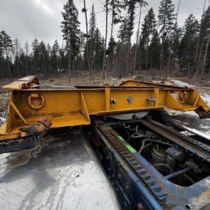 Abandoned yellow steel frame resting on a blue vehicle chassis in a snowy pine forest.