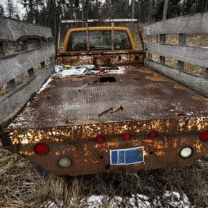 Rear view of a rusted flatbed trailer with wooden side rails, red taillights, and snow on the ground.