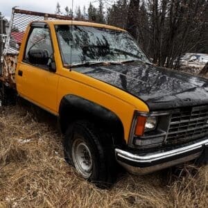 Old yellow and black pickup truck with a rusty flatbed sits in dry grass in a snow-dusted, rural lot. pieces of wood behind a fence in the background.