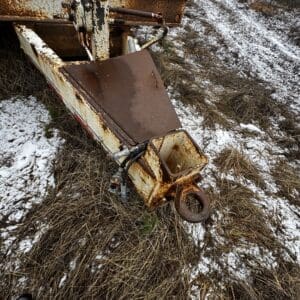 Rusty, abandoned industrial machine part with a large circular hitch sits on snow-dusted grass in a rural area.