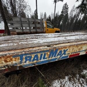 Rusty TRAILMAX flatbed trailer in a snowy, forested yard with a yellow truck cab in the background