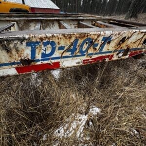 Abandoned, rusted boat hull with peeling blue and red graphics in a dry grassy field near a forest. Snow patches around.