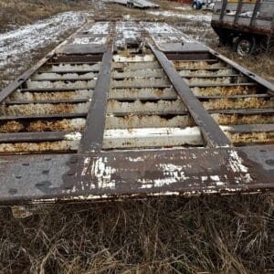 Rusty, open-frame flatbed trailer lying in a dry, snow-speckled lot with overgrown grass and a yellow vehicle in the background.