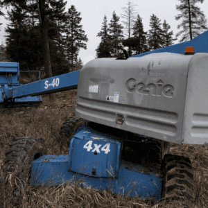 Blue Genie S-40 telescopic boom lift in a grassy, wooded field with its arm extended to the left; rugged tires and weathered equipment visible.