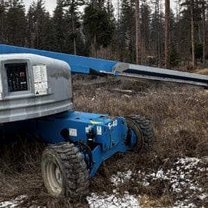 Blue Genie telescoping boom lift parked in a clearing with bare trees and patches of snow on the ground, ready for use in outdoor maintenance.