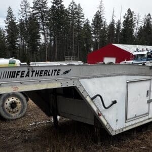 Featherlite aluminum flatbed trailer connected to a truck in a rural yard with pine trees and a red building nearby.