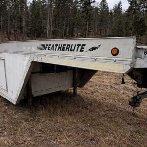 White Featherlite aluminum trailer frame on a dry grassy field with a forest backdrop, parked and unused.