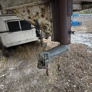 Rusty gooseneck trailer hitch under a raised frame, with chains and a ball, on a dry grassy lot.