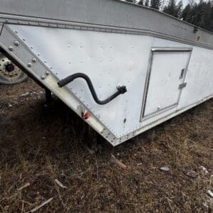 Aluminum cargo trailer side with a black curved hose/pipe attached, sitting on dirt with trees in the background.