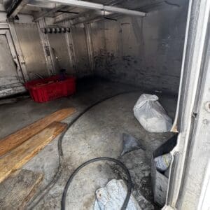 Interior of a dirty empty truck trailer with wooden planks, a red crate, hoses, and bags on the floor