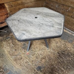 Weathered hexagonal outdoor table with a central hole for an umbrella, resting in a shallow dirt pit against wooden walls; dusty and dirty surface with straw on the ground.
