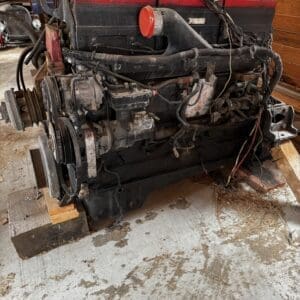 Large automobile engine block on a stand in a workshop, with belts, hoses and wiring visible and dusty/constrained setup.