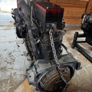 Rotary engine on a wooden stand in a dusty workshop with a red-top battery and exposed wiring attached to the engine block