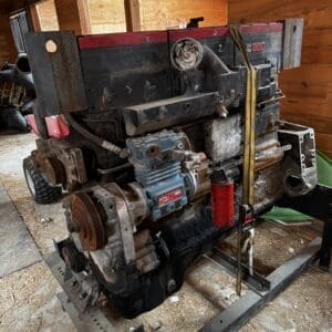 Diesel engine mounted on a metal frame in a wooden shed, showing rust, belts, hoses, and a blue pump component with a red cylindrical canister nearby.