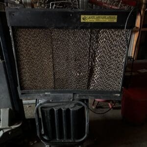 Old portable space heater with metal radiator fins in a cluttered workshop, yellow caution label on top.