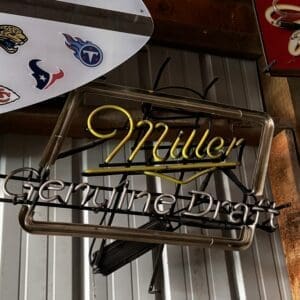 Neon sign reading 'Miller' with 'Genuine Draft' beneath, hanging on a metal wall beside a decorated surfboard with team logos.