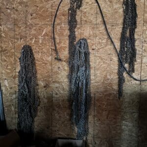 Chains hanging on a plywood-walled workshop with a black hose looped over a rail above, and a cardboard box of metal parts at the bottom left.
