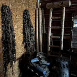 Chain bundles hang on a plywood wall beside stacked wooden ladders and weathered lumber in a cluttered workshop corner.