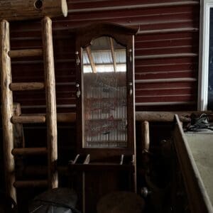 Old wooden armoire with a mirrored glass door, leaning against a red barn wall, surrounded by rustic logs and metal containers.