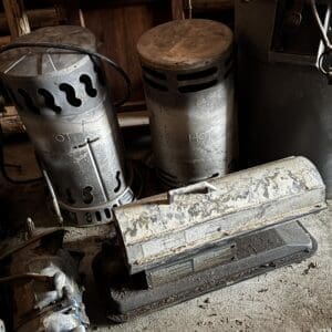 Three vintage metal kerosene heaters in a dusty storage space, with 'HOT' markings on two cylinders and a worn, rusted heater on the floor behind them.