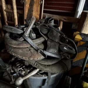 Dusty pile of worn leather horse saddles and reins atop a metal trash can in a cluttered shed.