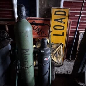Two chained gas cylinders (green and black) in a dim storage shed, with a yellow 'LOAD' sign leaning against a red metal wall behind them.