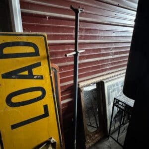 Yellow sign with black letters leaning against a red corrugated wall, next to a tall adjustable metal stand and several framed mirrors on the floor in a cluttered storage space.