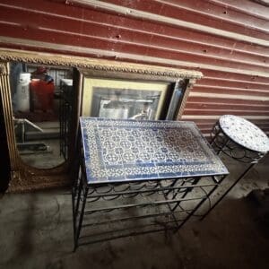 Antique-style storage scene with a gold-framed ornate mirror, framed art, and blue-and-white mosaic top tables against a red wooden wall in a dusty space.