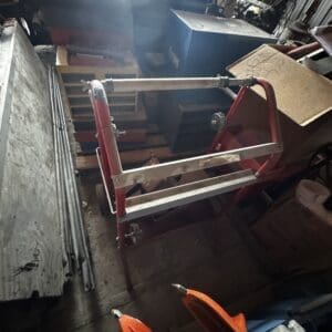 Red metal rolling cart with a metal pipe on top in a cluttered workshop, surrounded by boxes and tools nearby.