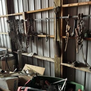 Rusty hand tools hang on a wooden rack inside a metal shed, with a red toolbox and boxes on the floor.
