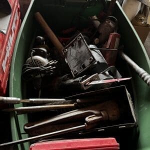 Green tool bin filled with dusty hand tools, saw blades, files, and a red plastic case.