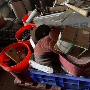 Cluttered workshop with stacked sanding discs, metal scraps, and tools in red buckets and a blue crate on a pallet.