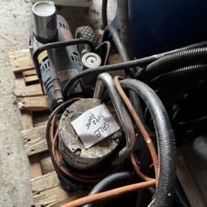 Close-up of a dusty electric motor/pump with tangled orange and black cables, sitting on a wooden pallet in a cluttered workshop. A handwritten note is taped to the device.